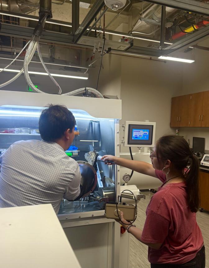 Prof. Vladislav Klepov assists a student in measuring the radiation of uranium compounds. Image shows male professor and female student standing front of a fume hood with a lab instrument next to it.