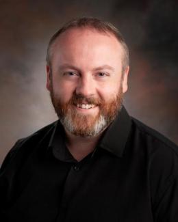 Image depicts Dr. Christopher Barrett, bearded caucasian man in a black shirt