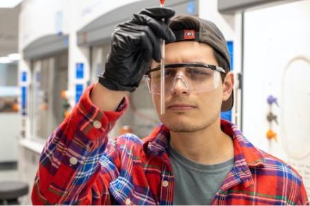 Image depicts young man in a lab, peering at the contents of a tube